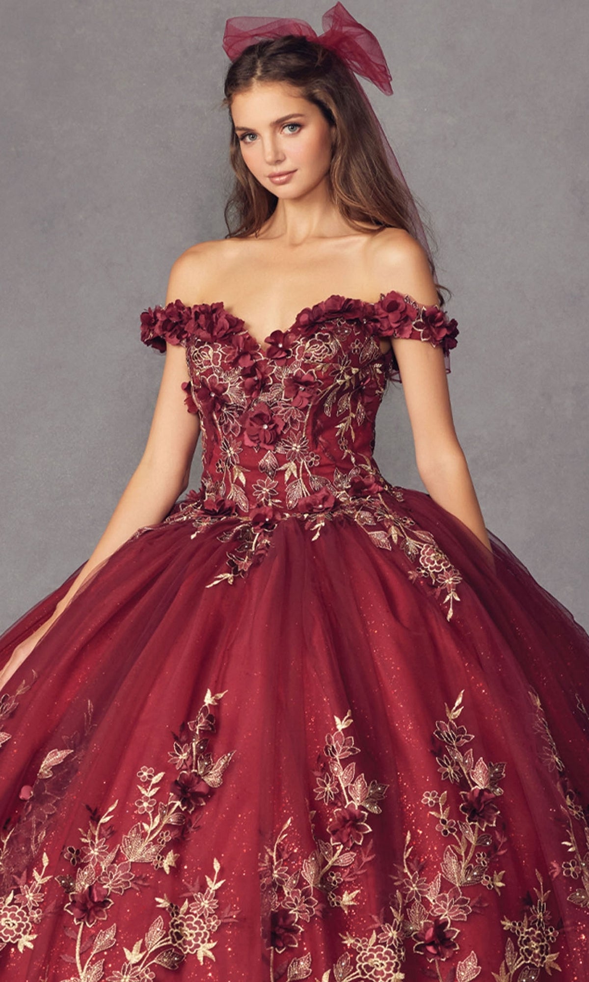 Woman poses against a gray background wearing the Juliet 1447 Off-the-Shoulder Quinceañera Dress in burgundy with beaded embroidery and a matching hair bow.